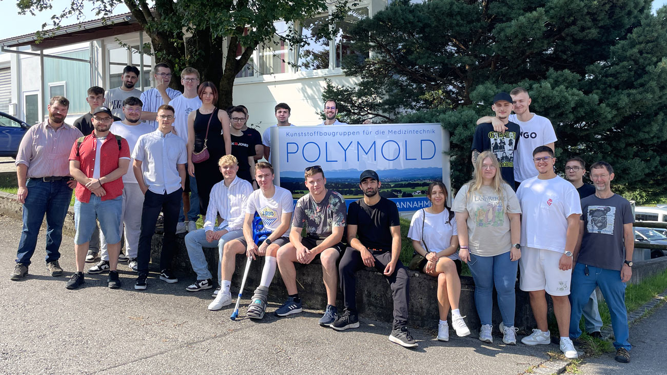 Werkzeugmacher-Berufsschulklasse aus Weilheim zu Besuch bei POLYMOLD | Gruppenbild zum Abschluss vor dem Firmengebäude in Pähl, Inhaber Jonathan Welz hinter dem Firmenschild über dem "L" zu sehen